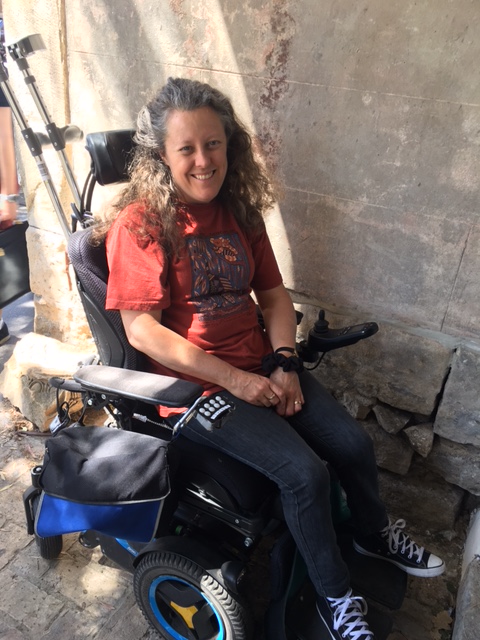 Jane is sitting in her power wheelchair, smiling. She has long curly brown hair, and is wearing a rust coloured t-shirt, jeans and gym-boots. She has crutches behind her. She's next to a sandstone wall.