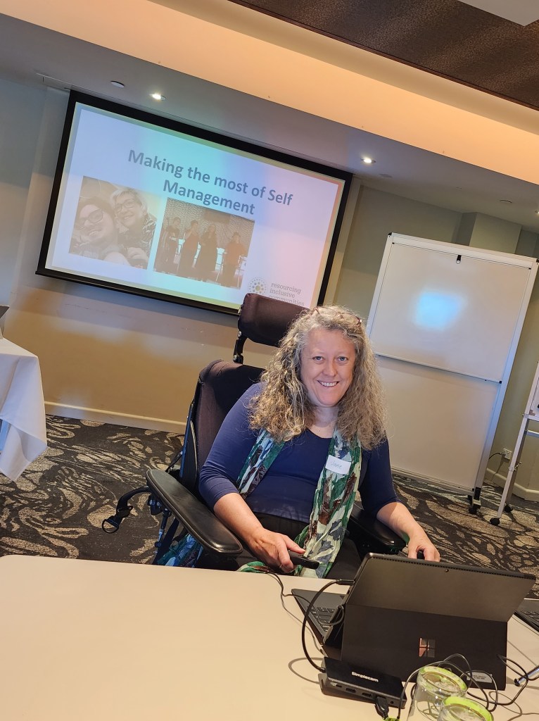 Jane in a wheelchair and business clothes, in front of a screen, in a conference room.