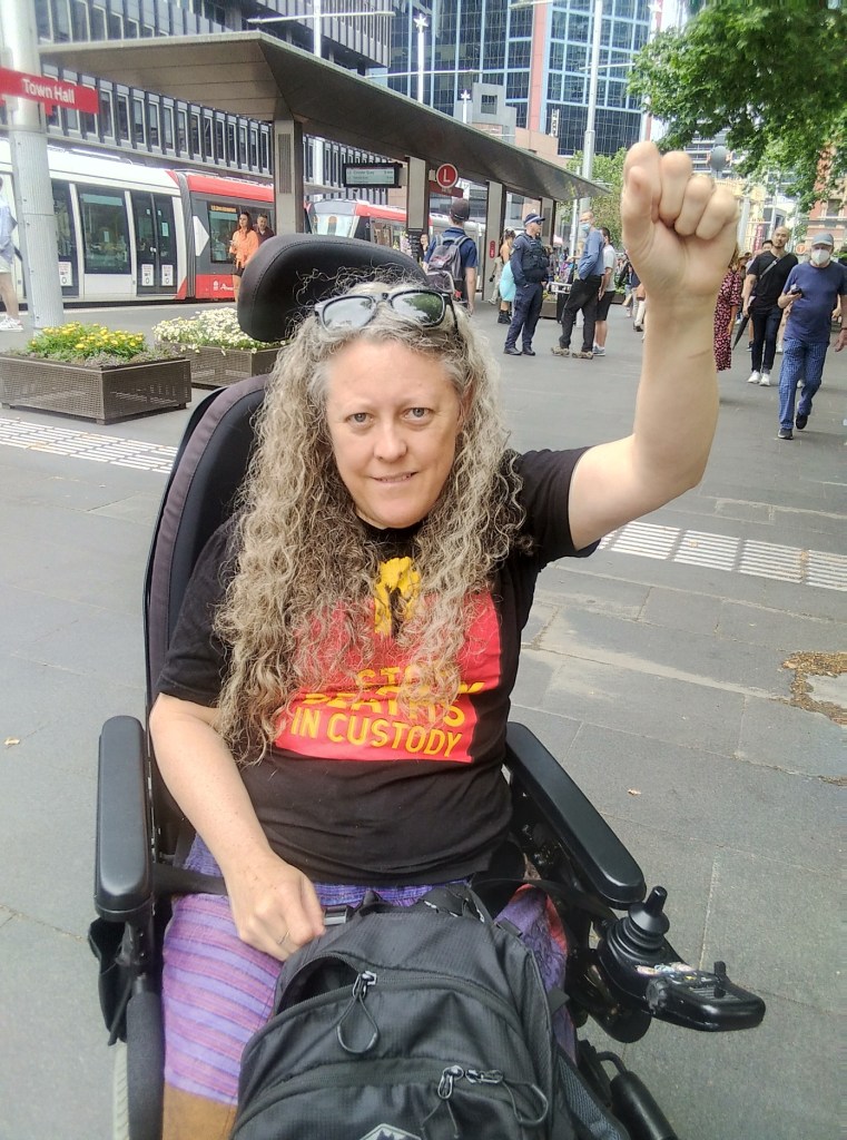 Jane in her wheelchair, wearing Stop Black Deaths in Custody shirt, fist raised.
