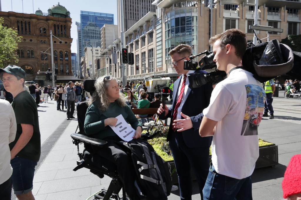 Jane in wheelchair, outdoors, being interviewed by a journalist and filmed by a camera man.