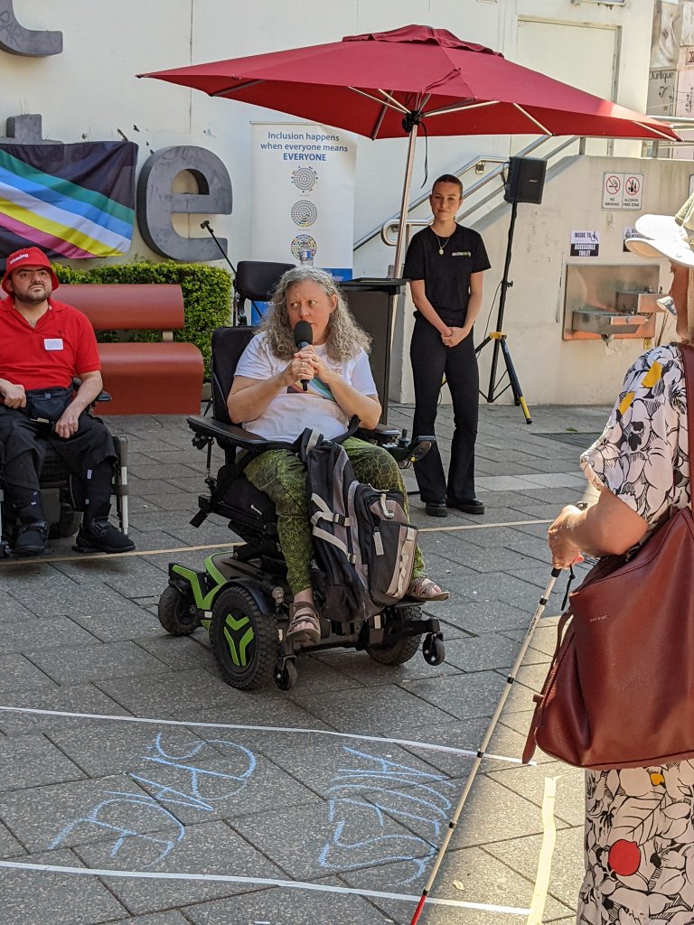 Jane in a wheelchair, outdoors, speaking into a microphone with the disability pride flag in the background.