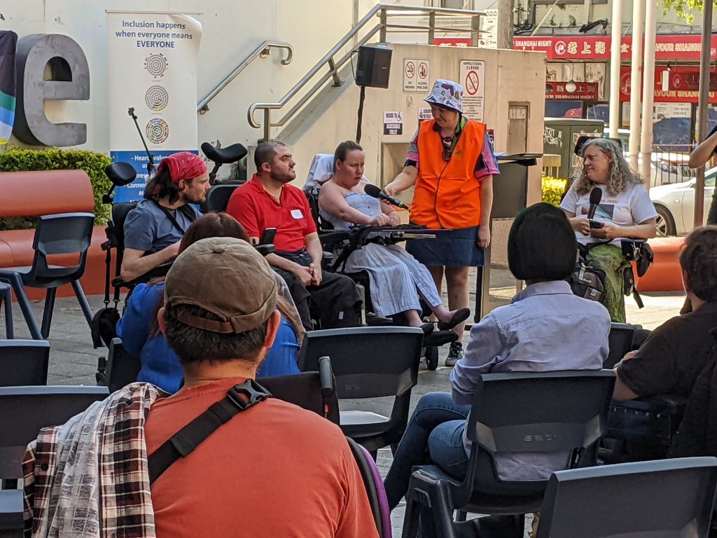 Four wheelchair users on an outdoor stage, in front of an audience