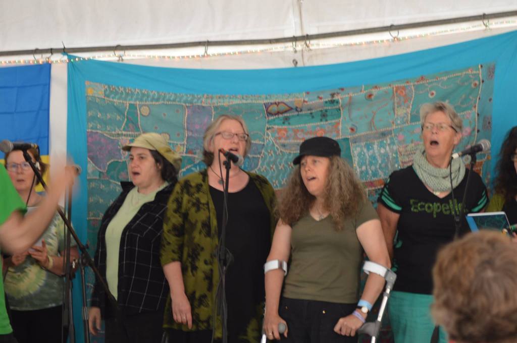 Five women, dressed in green and black singing on stage into microphones.