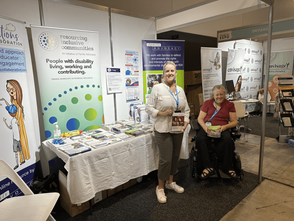 Jane, in her wheelchair, Jacquie standing in front of banners and an info table.