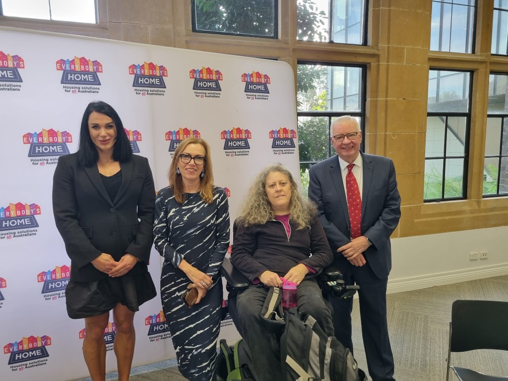 Four people in front of the Everybody's Home banner. Jane in a wheelchair.