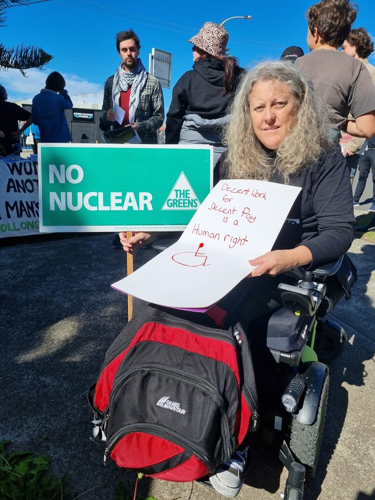 Jane at a rally, in wheelchair, with a "no nuclear" sign and one calling for fair pay