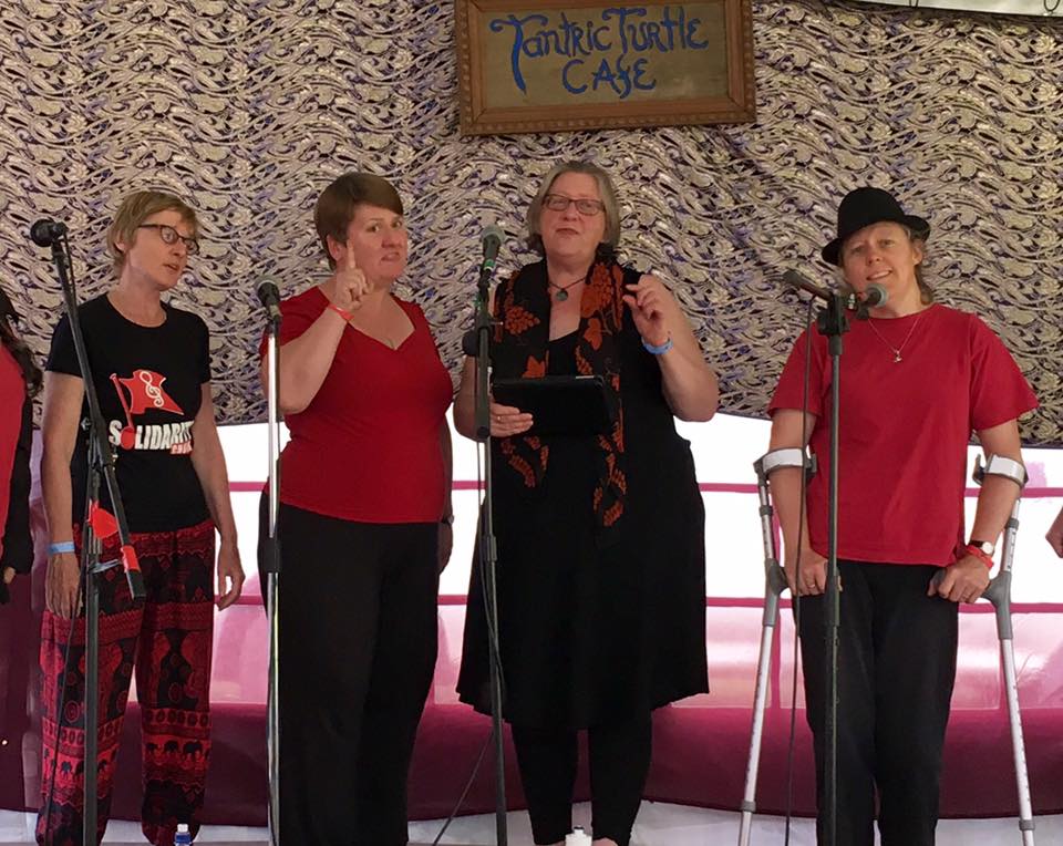 Four women in red and black singing into microphones. One points to the audience with eyebrows raised.