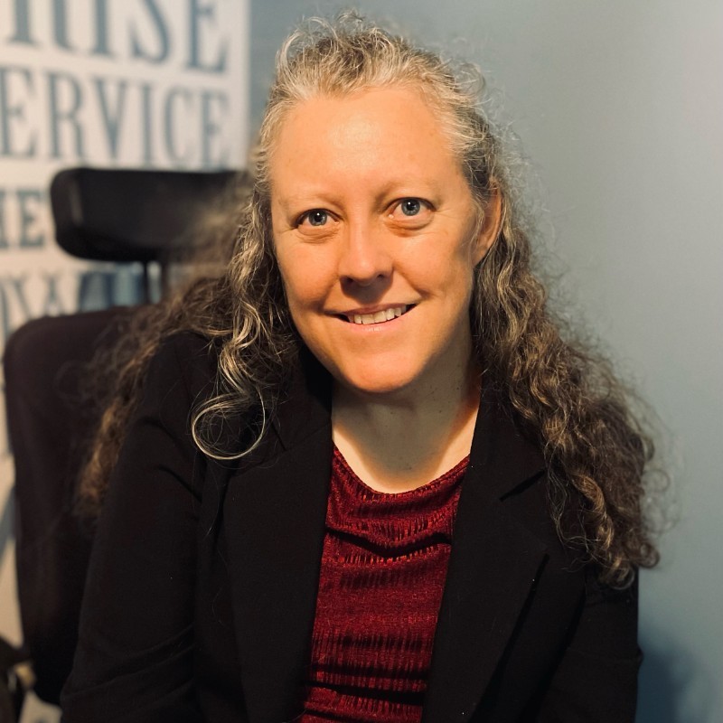 Smiling white woman with long curly brown hair, in business clothes, sitting in a wheelchair
